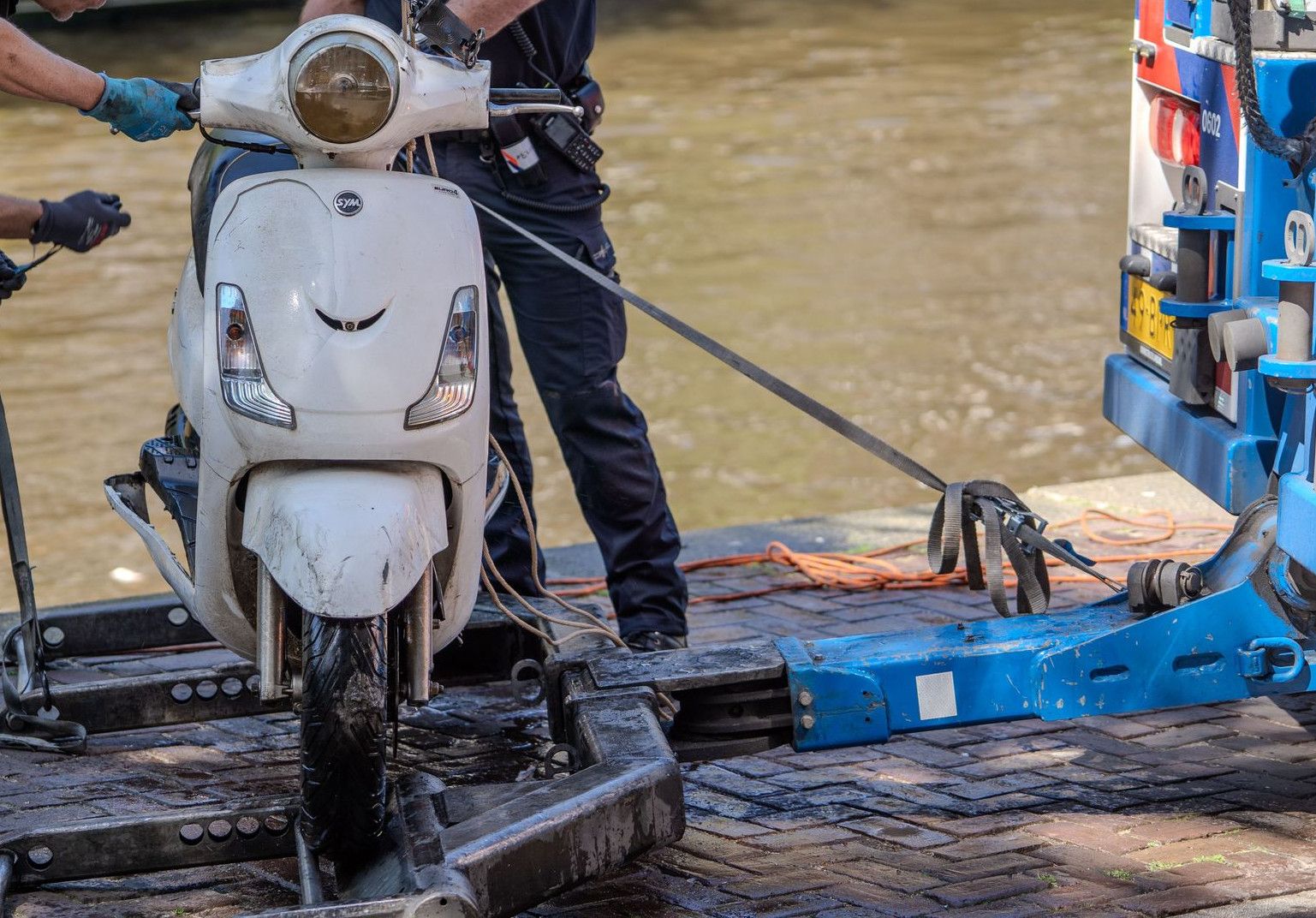 Police officers retrieve a motor scooter from a canal using towing equipment.